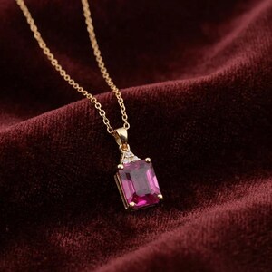 May include: A gold necklace featuring a rectangular emerald-cut magenta gemstone pendant. The pendant is accented with three small clear stones at the top where it connects to a delicate gold chain, creating a refined and elegant jewelry piece.