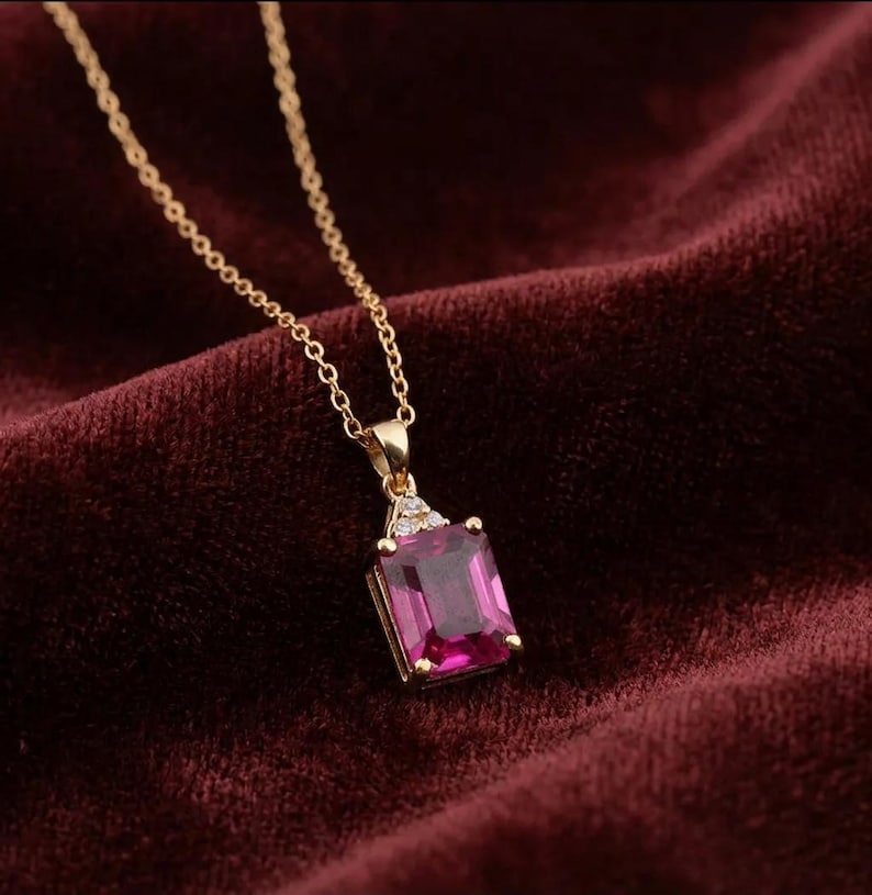 May include: A gold necklace featuring a rectangular emerald-cut magenta gemstone pendant. The pendant is accented with three small clear stones at the top where it connects to a delicate gold chain, creating a refined and elegant jewelry piece.