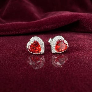 May include: A pair of stud earrings featuring a central heart-shaped red gemstone surrounded by a halo of small clear stones set in a silver-toned metal frame. The earrings are positioned side by side, displaying a reflective surface finish.