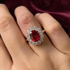 May include: A ring featuring a large, oval-cut deep red gemstone at the center, surrounded by a halo of twelve round, clear, faceted stones. The setting is a metallic band that holds the central cluster securely in place on the finger.
