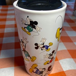 May include: White travel mug with a black lid featuring multiple cartoon images of Mickey Mouse. The mug has a cylindrical shape and is on a red and orange checkered tablecloth. The mug is approximately 15 cm tall.