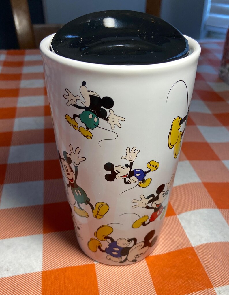 May include: White travel mug with a black lid featuring multiple cartoon images of Mickey Mouse. The mug has a cylindrical shape and is on a red and orange checkered tablecloth. The mug is approximately 15 cm tall.