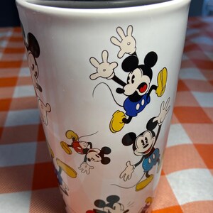 May include: White travel mug decorated with multiple images of Mickey Mouse in various poses. Mickey is shown in his classic outfit with red shorts, yellow shoes, and white gloves. The mug has a black lid and is on a checkered orange and white surface.
