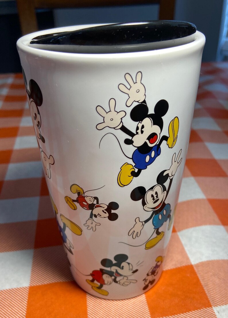 May include: White travel mug decorated with multiple images of Mickey Mouse in various poses. Mickey is shown in his classic outfit with red shorts, yellow shoes, and white gloves. The mug has a black lid and is on a checkered orange and white surface.
