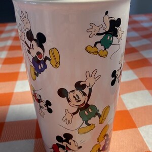 May include: White travel mug decorated with multiple images of Mickey Mouse in different poses. Mickey is shown with his black ears, red shorts, yellow shoes, and white gloves. The mug has a black lid and is cylindrical in shape.