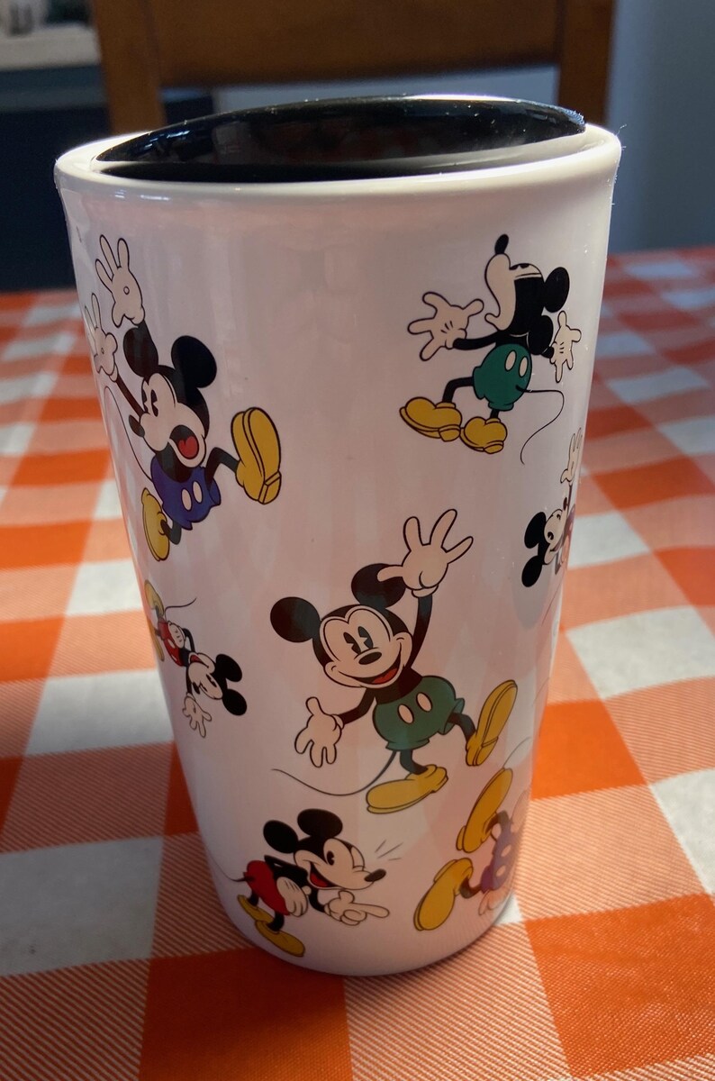 May include: White travel mug decorated with multiple images of Mickey Mouse in different poses. Mickey is shown with his black ears, red shorts, yellow shoes, and white gloves. The mug has a black lid and is cylindrical in shape.