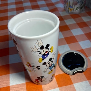 May include: White ceramic travel mug decorated with Mickey Mouse illustrations. The mug includes a black lid with Mickey Mouse ear detailing. The cylindrical mug is empty. The background is a red and white checkered pattern.