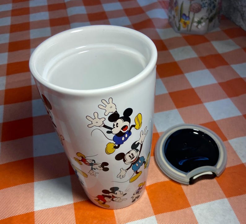 May include: White ceramic travel mug decorated with Mickey Mouse illustrations. The mug includes a black lid with Mickey Mouse ear detailing. The cylindrical mug is empty. The background is a red and white checkered pattern.