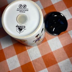 May include: White ceramic Disney travel mug with a black lid. The mug features images of Mickey Mouse and other Disney characters. The bottom of the mug has the Disney logo and text that reads "MADE IN THAILAND".