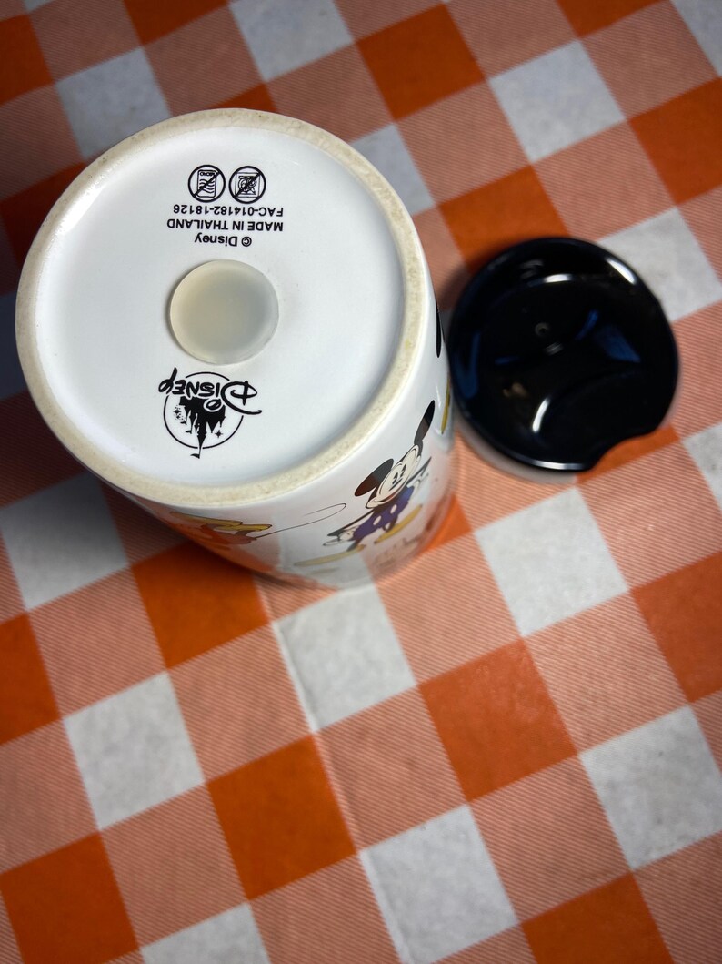 May include: White ceramic Disney travel mug with a black lid. The mug features images of Mickey Mouse and other Disney characters. The bottom of the mug has the Disney logo and text that reads "MADE IN THAILAND".