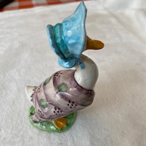 May include: A ceramic duck figurine wearing a light blue bonnet with a ruffled edge. The duck has a white neck and a purple body decorated with green polka dots and small black markings. It stands on a small green base with orange feet.
