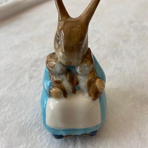 May include: A ceramic figurine of a brown rabbit wearing a blue coat and a white apron, holding two smaller brown and white baby rabbits. The rabbit has long, upright ears and a glossy finish, with painted facial features and detailed fur textures.