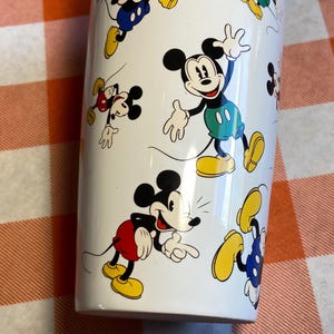May include: White cylindrical tumbler decorated with cartoon images of Mickey Mouse. Mickey is shown in different poses, wearing his iconic red shorts, yellow shoes, and white gloves. The tumbler is white.
