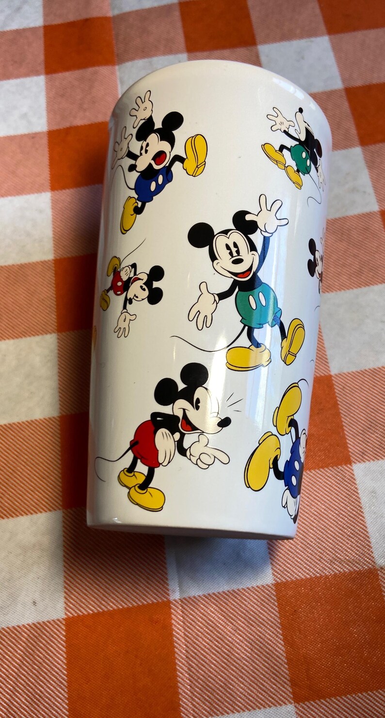 May include: White cylindrical tumbler decorated with cartoon images of Mickey Mouse. Mickey is shown in different poses, wearing his iconic red shorts, yellow shoes, and white gloves. The tumbler is white.