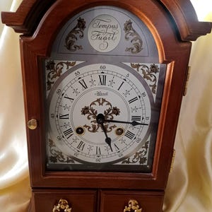 May include: A wooden mantel clock with a dark brown finish, featuring a white face with black Roman numerals, the text Tempus Fugit, and the brand name Hermle. The clock includes black hands, gold-toned hardware, and two small drawers at the base.