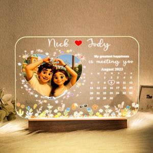 May include: A personalized LED night light with a photo of a couple inside a heart-shaped frame. The light includes the names "Nick" and "Judy", the text "My greatest happiness is meeting you", and a calendar for August 2023. The base is made of wood.