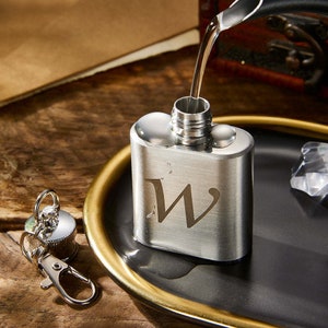 Op de afbeelding: Een zilveren fles met een sleutelhanger eraan. De fles heeft een gegraveerde letter "W". De fles wordt gevuld met vloeistof.