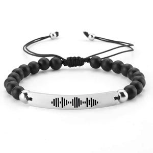 Puede incluir: Pulsera de cuentas negras con cierre ajustable plateado y una placa plateada con un gráfico de onda sonora.