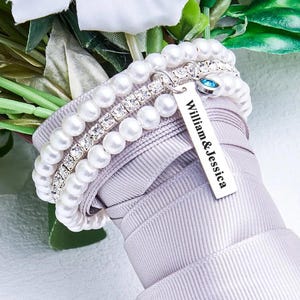 Op de afbeelding: Een armband met meerdere strengen, bestaande uit twee rijen witte kunstparels en een centrale rij met heldere strass-steentjes. Een zilverkleurige rechthoekige metalen bedel is gegraveerd met de tekst William & Jessica en een blauw kristal.