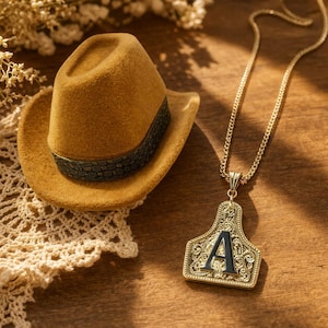 May include: Mustard-colored cowboy hat with a patterned band and a gold-colored necklace with a pendant. The pendant has a black letter "A" and ornate detailing. The items are on a wooden surface.