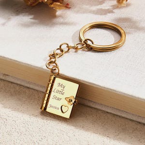 May include: A gold-coloured keychain with a small book pendant. The book is inscribed with "My Little Star" and the name "Jessica", plus a heart symbol. The keychain is connected to a large ring, suitable for keys or a bag charm.