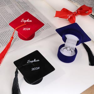 Könnte beinhalten: Zwei Ringboxen in Form von Abschlusskappen. Eine ist aus rotem Samt mit dem Namen "Sophia" und dem Jahr "2024" bestickt. Die andere ist aus schwarzem Samt mit der gleichen Stickerei. Beide Boxen haben einen Quasten.