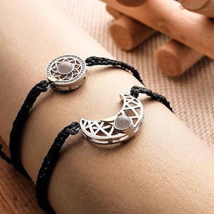 Puede incluir: Dos pulseras plateadas a juego, una con un diseño de sol y la otra con forma de media luna. Ambas tienen una piedra central y están unidas por cordones negros. Un accesorio con temática celestial.