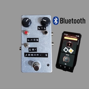 Op de afbeelding: Een zilveren gitaareffectpedaal met zwarte knoppen, een rode knop en de tekst "KNOB EX' JOHNN. 5". Een smartphone toont een besturingsinterface, verbonden via Bluetooth. Het Bluetooth-symbool en de tekst zijn zichtbaar.