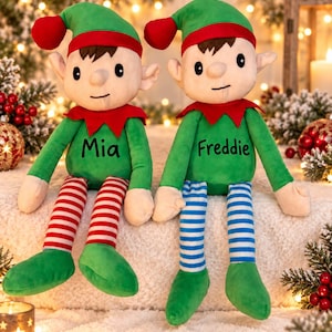 May include: Two plush elf dolls with green outfits, red collars, and pointed hats with red pom-poms. One elf has red and white striped legs and the name Mia embroidered on its chest. The other has blue and white striped legs and the name Freddie.
