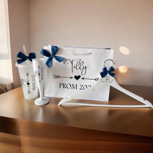 May include: A white gift bag with a blue bow and the text "Tilly Prom 2023" printed on it. A white hanger with a blue bow and the text "Tilly Prom 2023" printed on it. A clear plastic cup with a blue bow and the text "Tilly Prom 2023" printed on it.