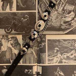 Puede incluir: Cinturón de cuero negro con hebilla y adornos decorativos de metal plateado. El cinturón presenta una serie de adornos circulares y ovalados, junto con un detalle de cadena. La hebilla y los adornos tienen un diseño grabado. El cinturón se muestra en una página de revista vintage.