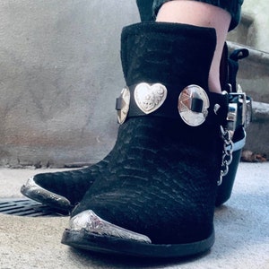 May include: Black suede ankle boots with silver-tone metal accents. The boots feature decorative metal heart and star-shaped embellishments on a black leather strap around the ankle. Silver metal is also on the toe.