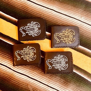 May include: Four dark brown leather wallets with a white or gold scorpion design. The wallets are arranged on a striped blanket. Each wallet has a stitched border.