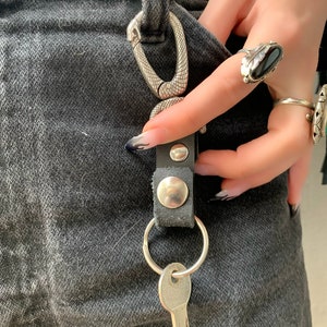 Op de afbeelding: Een zilveren sleutelring met een sleutel eraan, vastgehouden door een zwarte leren riem met zilveren klinknagels. De sleutelring is verbonden met een zilveren karabijnhaak met een slangenhuidpatroon.