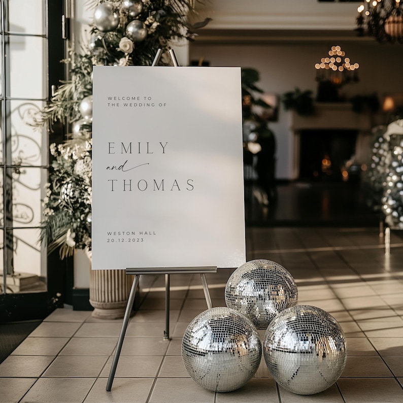 May include: A white wedding sign with black text that reads "Welcome to the wedding of Emily and Thomas, Weston Hall, 20.12.2023". The sign is on a black easel and there are three silver disco balls in front of it.