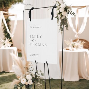 May include: A white wedding welcome sign with black text that reads "Welcome to the wedding of Emily and Thomas, Weston Hall, 20.12.2023". The sign is on a black easel with white flowers and greenery on the left and right sides.
