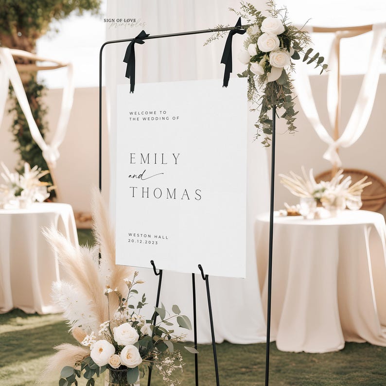 May include: A white wedding welcome sign with black text that reads "Welcome to the wedding of Emily and Thomas, Weston Hall, 20.12.2023". The sign is on a black easel with white flowers and greenery on the left and right sides.
