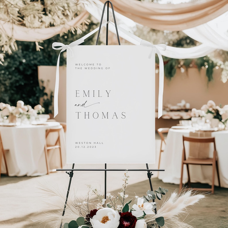May include: White wedding sign with black text that reads "Welcome to the wedding of Emily and Thomas, Weston Hall, 20.12.2023". The sign is on a black easel with white ribbon.