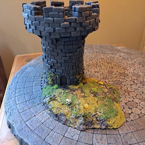May include: A miniature stone tower with a small door and a grassy base, set on a circular stone base with a cobblestone pattern.
