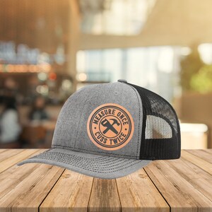 May include: A gray and black trucker hat with a brown leather patch that says "Measure Once Cuss Twice" with a hammer and saw graphic.