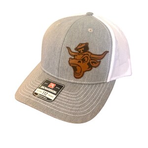 May include: A gray and white trucker hat with a brown leather bull's head patch. The patch features a bull with a cowboy hat on its head. The hat is adjustable and has the text "Richardson 112 Trucker" on the side.