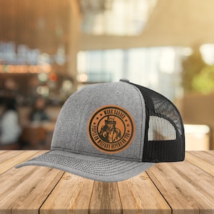 Puede incluir: Una gorra de camionero gris y negra con un parche de cuero marrón que presenta un dibujo animado del Tío Sam con el texto "Work Harder People on Welfare Depend on You".