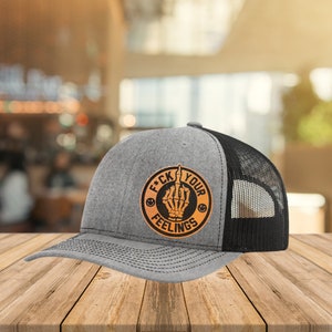 Op de afbeelding: Een grijze en zwarte truckercap met een bruin leren patch waarop "F*CK YOUR FEELINGS" staat met een skelet-middelvinger.