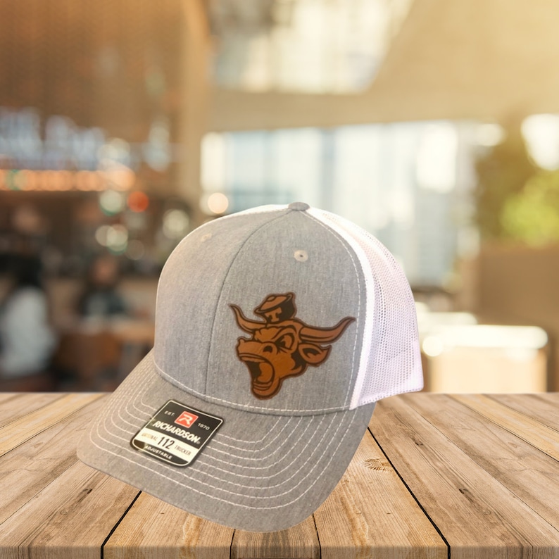 May include: A gray and white trucker hat with a brown leather bull logo. The logo features a bull with a cowboy hat on its head. The hat is adjustable and has a mesh back.