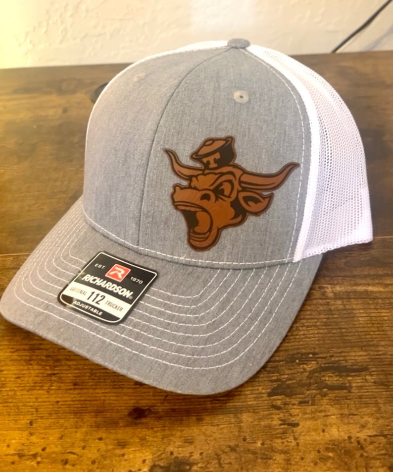 May include: Gray and white trucker hat with a brown leather patch of a bull wearing a cowboy hat. The patch is on the front of the hat. The hat has a white mesh back and a gray front. The hat has a Richardson 112 adjustable label.