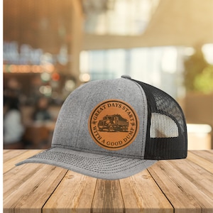 Peut inclure: Un chapeau de camionneur gris et noir avec un patch en cuir marron qui dit "Great Days Start With A Good Dump" et représente la silhouette d'un camion à benne.
