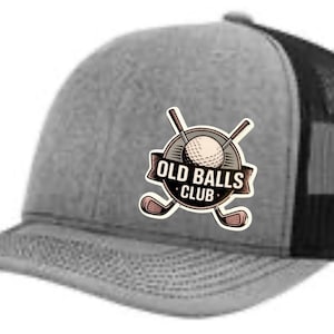 Könnte beinhalten: Grau-schwarze Trucker-Cap mit gebogenem Schirm. Das Cap hat ein rundes Emblem mit Golfball, gekreuzten Golfschlägern und dem Text "OLD BALLS CLUB". Die Rückseite ist aus Mesh.