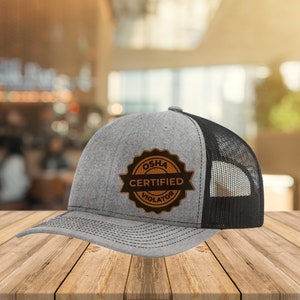 Könnte beinhalten: Eine graue und schwarze Trucker-Kappe mit einem braunen Leder-Patch, auf dem "OSHA Certified Violator" steht.
