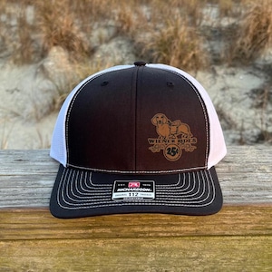 May include: Black and white trucker hat with a brown embroidered dachshund wearing a saddle and the text "Wiener Rides 25th" on the front. The hat is made by Richardson.
