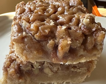 May include: A close-up of a pecan pie bar with a thick layer of pecan topping and a flaky crust. The bar is cut into two pieces, revealing the layers of the dessert.